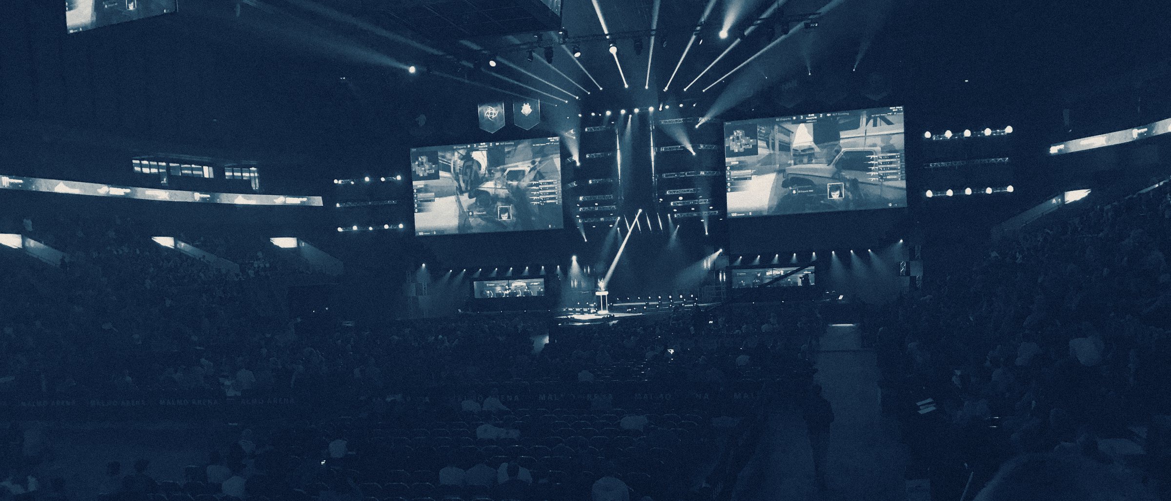 A packed esports arena during a live match, stage and screens lit above the crowd
